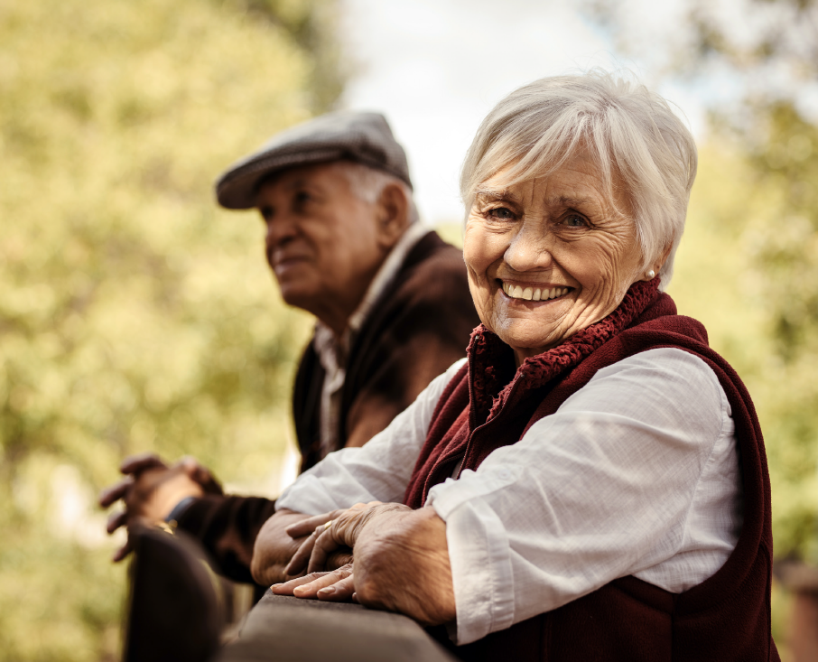 To ældre mennesker, der smiler. De er udenfor og står op af et hegn.